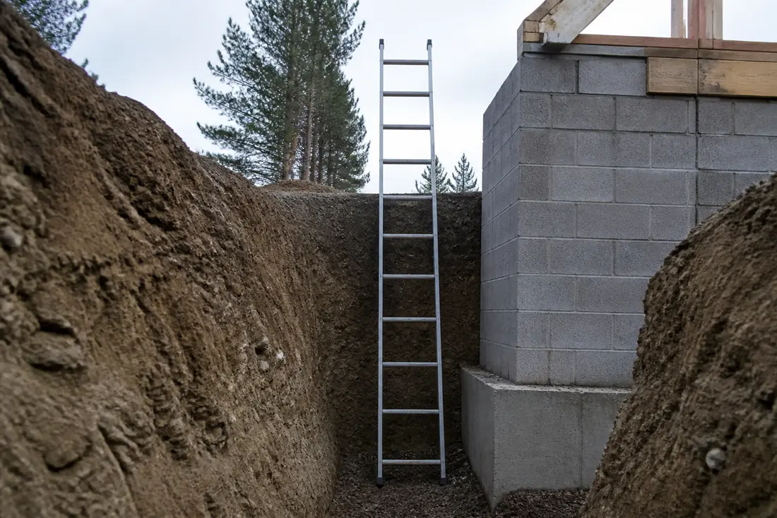 A ladder standing in an open trench beside a partly built wall, reaching above the top of the cut